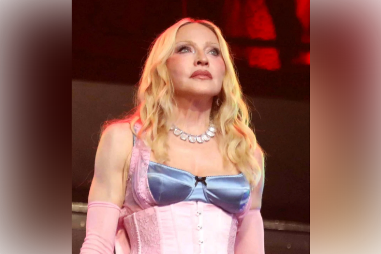 Madonna in a pink corset dress and gloves stands onstage under red lights, gazing upward with a calm, dramatic expression.