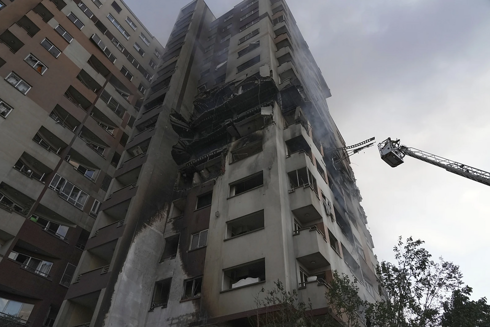 High-rise building with charred, smoking exterior from a fire. Firetruck ladder extends upward. Overcast sky and trees in the foreground.