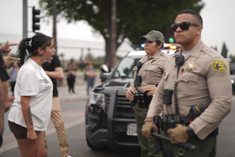 A woman in a white shirt confronts two uniformed officers in front of a police car. The scene is tense, set on a city street.