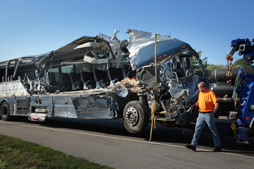 Damaged bus with a torn roof on the roadside. A man in an orange shirt walks next to it. Clear sky, trees in the background.