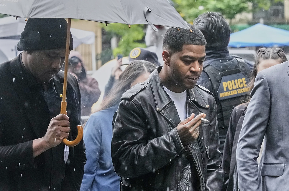 Kid Cudi in leather jacket smoking in rain. Another man holds umbrella. Police with "Homeland Security" vest in background. Crowded street.