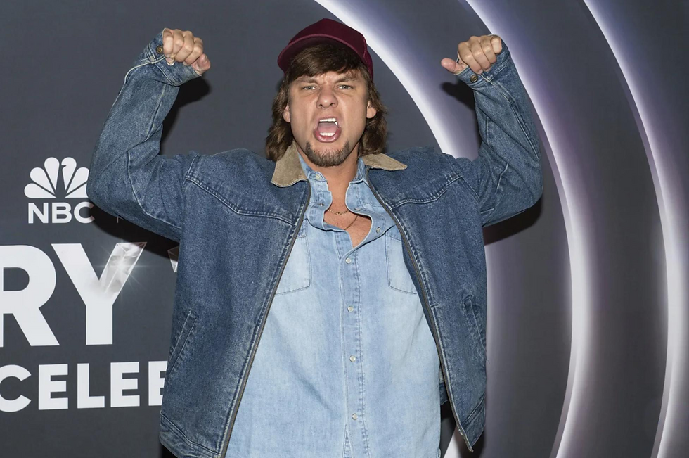 Man in a denim jacket and cap flexes arms, shouting energetically against a dark background with NBC logo and "RY" text.