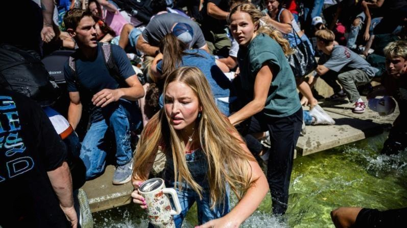 Crowd of young people urgently fleeing over water, midday, carrying personal items. Focused expressions, vibrant clothing, sunny setting.