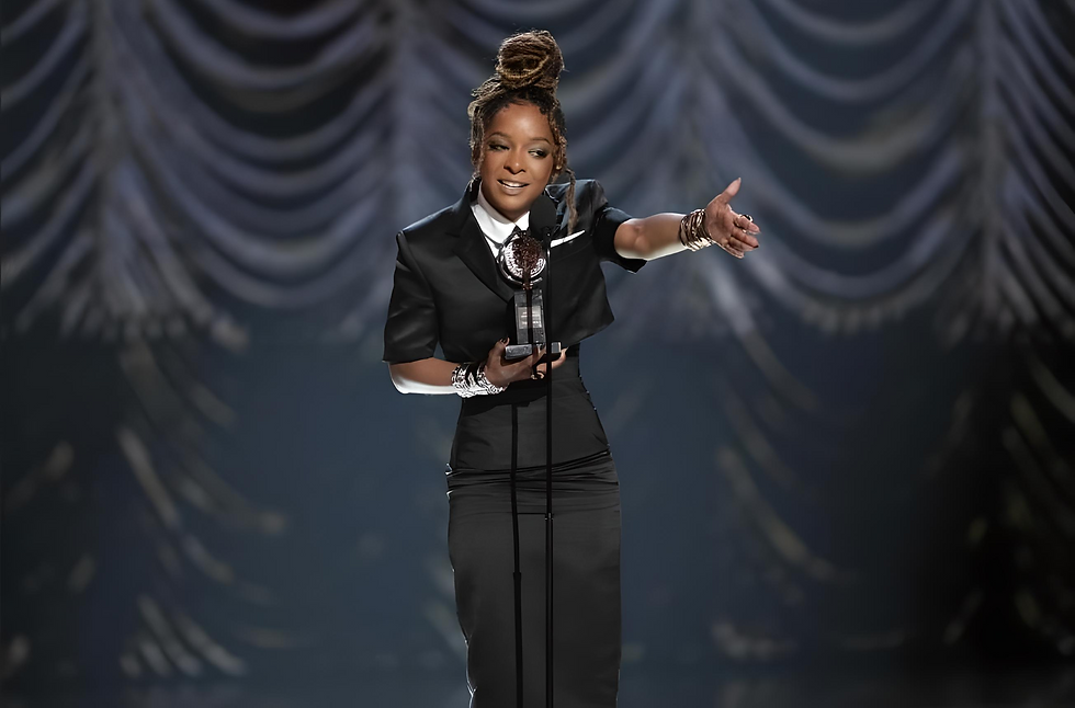 Kara Young in black attire holds an award onstage, gesturing with one hand. Background features draped fabric. Calm and confident mood.