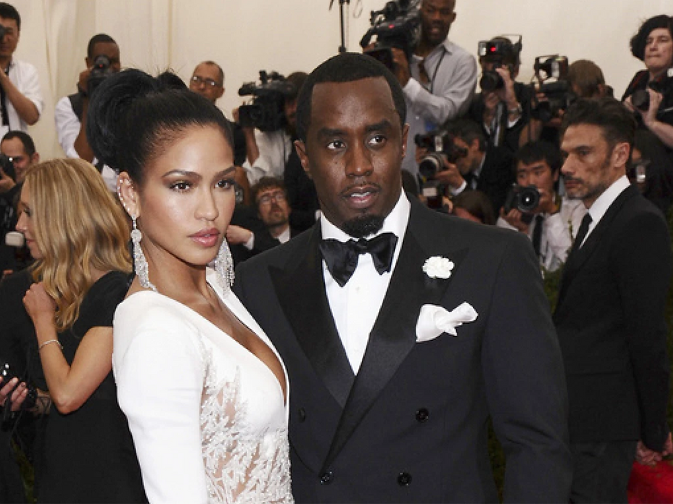 Couple in elegant attire pose on a red carpet, surrounded by photographers. She wears a white dress; he dons a black tuxedo with a white boutonniere.
