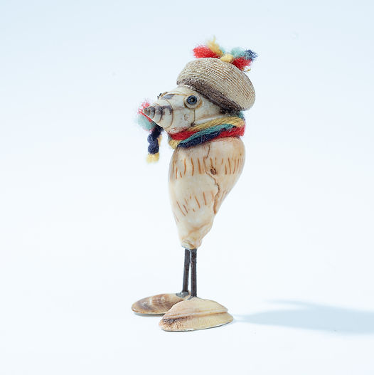 A smaller Shellfolk figure made from shells, standing upright with a shell hat and colourful scarf, appearing friendly and attentive.