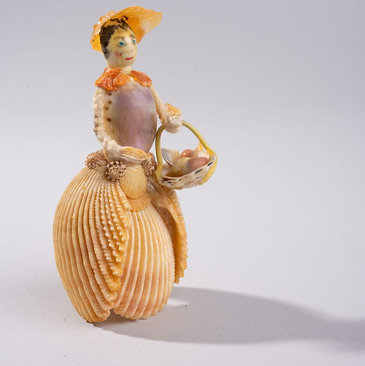 Shellfolk figure wearing a layered shell skirt and wide-brimmed shell hat, holding a small basket made from shells.