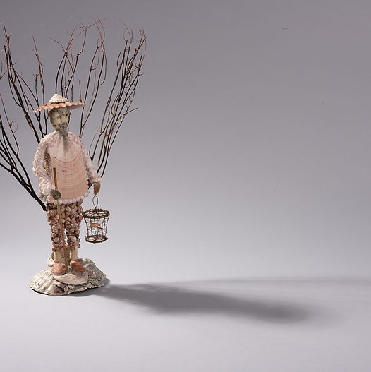 Shellfolk figure of Ling the Fisherman made from pale shells, wearing a shell hat, holding a wire cage with a small prawn inside, with branching sea twigs behind him on a shell base.