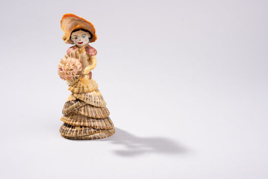 A standing Shellfolk figure made from layered shells, wearing a wide shell hat and holding coral flowers, with a gentle, calm expression.