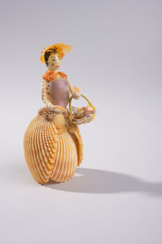 Shellfolk figure wearing a layered shell skirt and wide-brimmed shell hat, holding a small basket made from shells.