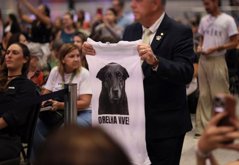 Em um evento oficial com público sentado ao fundo, um homem vestindo terno escuro, camisa branca e gravata clara segura uma camiseta branca em frente ao corpo. A camiseta traz estampada a foto em preto e branco de um cachorro preto com uma auréola desenhada sobre a cabeça, acompanhada do texto em letras garrafais "ORELHA VIVE!".
