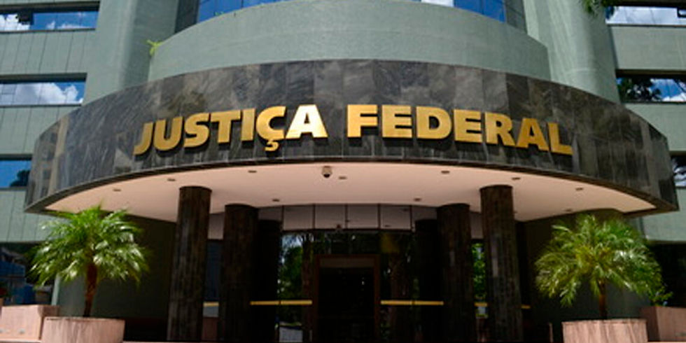 Fachada da entrada de um prédio público com uma estrutura arqueada de mármore escuro acima da recepção, exibindo a inscrição em grandes letras douradas "JUSTIÇA FEDERAL". A entrada conta com colunas cilíndricas escuras e dois vasos com pequenas palmeiras decorativas nas laterais, sob uma iluminação embutida no teto.