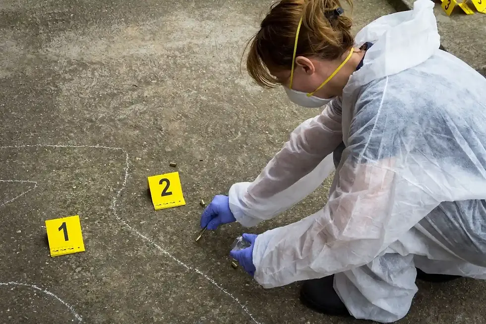 Uma perita criminal vestindo macacão impermeável branco de proteção, máscara facial descartável e luvas cirúrgicas azuis trabalha agachada em uma cena de crime ao ar livre. Ela utiliza uma pinça para coletar cápsulas de bala deflagradas do chão de concreto, posicionando marcadores de evidência numéricos amarelos com os números "1" e "2" ao lado de uma silhueta desenhada com giz branco.