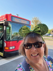 Woman in green Mickey ears smiles in front of a red bus labeled "Magic Kingdom." Bright day with blue sky and trees in background.