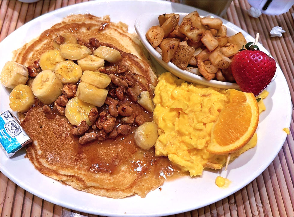 Plate of pancakes with bananas and nuts, scrambled eggs, diced potatoes, strawberry, and orange slice on a bamboo mat. Packs labeled "Butter." This is the banana fosters pancakes breakfast at Rainforest Cafe at Disney's Animal Kingdom Theme Park.