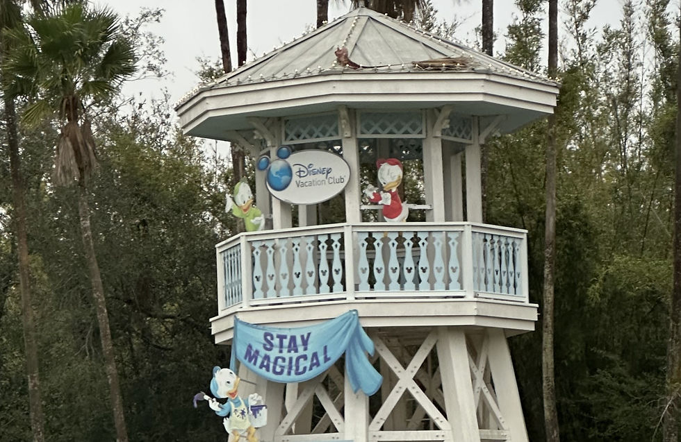White wooden tower with Disney Vacation Club sign, cartoon characters, and a "Stay Magical" banner. Palm trees and greenery in the background.