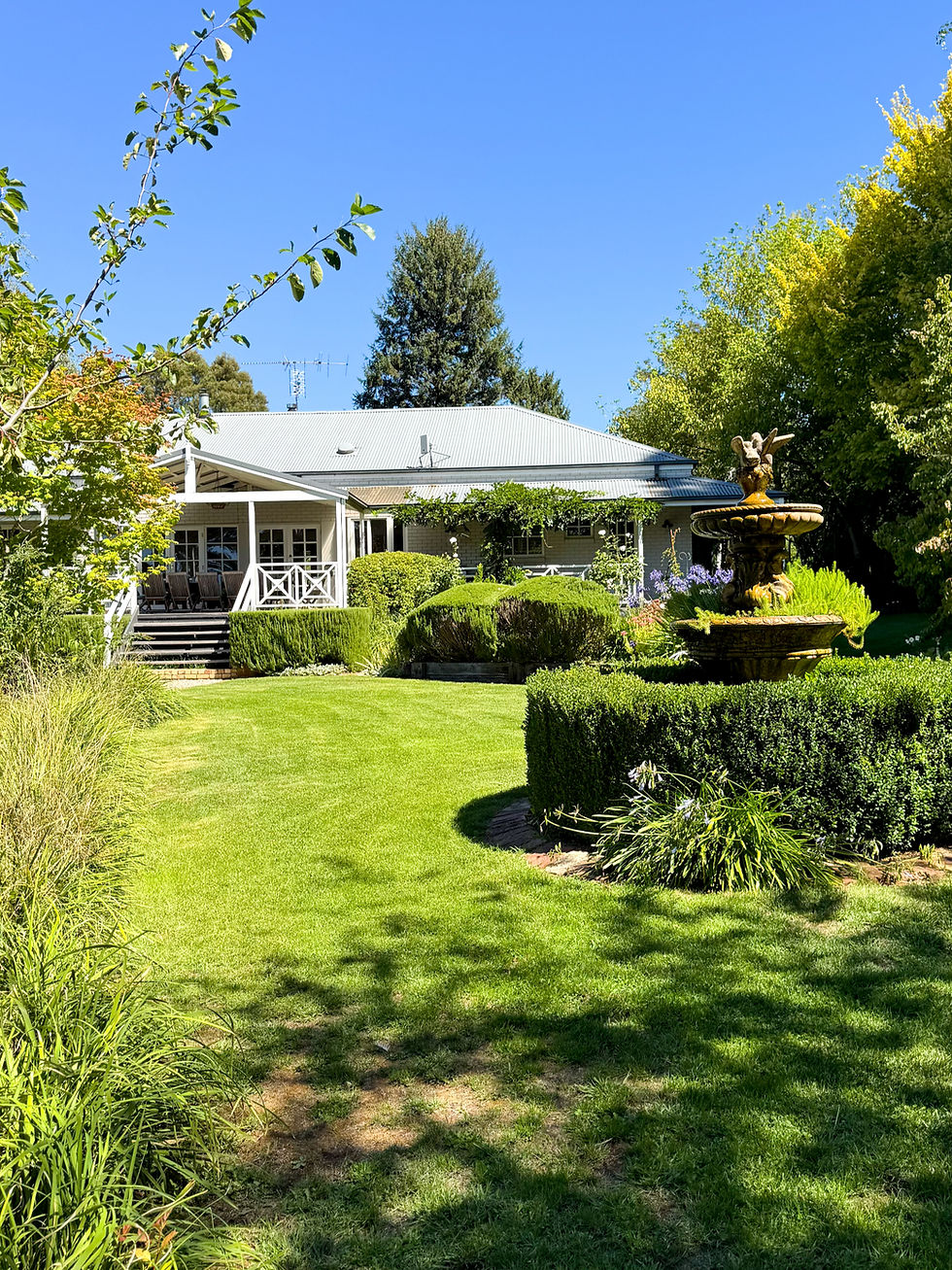A house with a porch and lush garden, featuring a stone fountain. Blue sky and green trees in the background create a serene atmosphere.