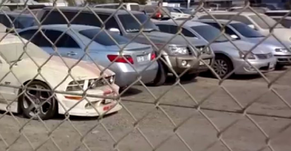 Cars parked behind a chain-link fence in a lot. A dusty car is in the foreground. The background shows more vehicles and a blue sky.