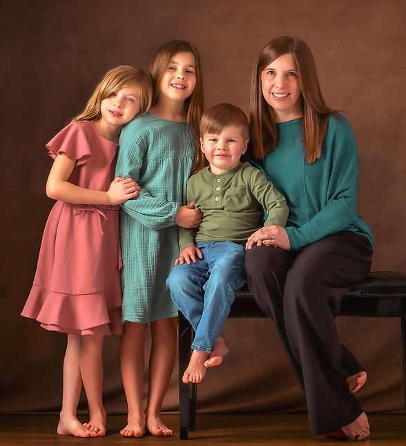 A working mom with her three children, two girls and a toddler boy wearing linen cotton pastels. The mother is sitting on a bench wearing a teal sweater, with her little guy next to her wearing a green shirt and denim with his biggest sister standing right next to him wearing a green dress and holding his hand, while her little sister is right next to her leaning her head lovingly on her for a motherhood and personal branding session for a therapist at the portrait by Taru Studio, a family and women photographer in Plymouth Minnesota MN Midwest