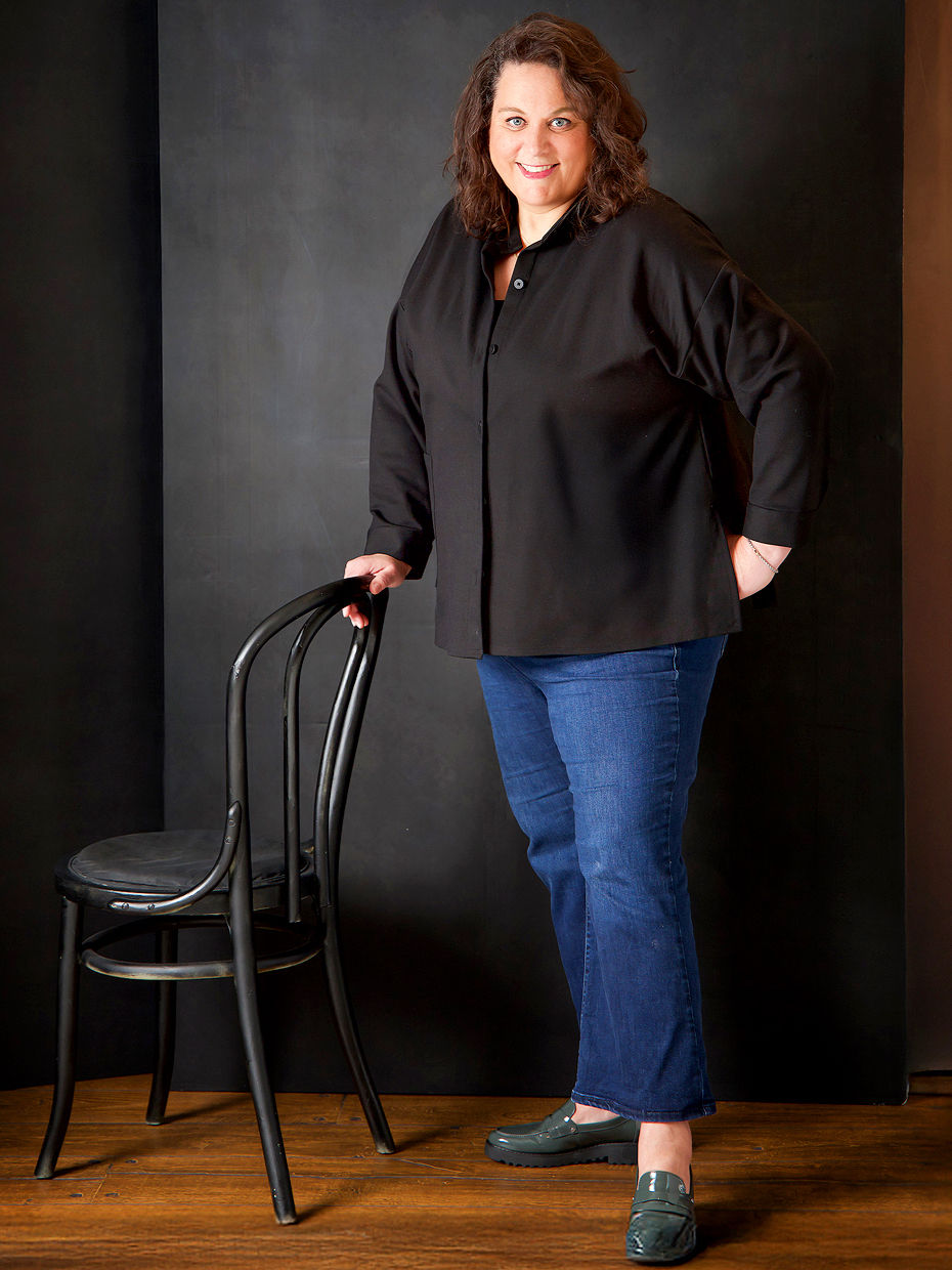 Personal branding corporate leadership headshot photography session for a curvy woman who in the testimonial said that she is so thankful that it’s not AI. She is standing with confidence, wearing a blue jeans and black jacket with one hand in pocket and other on the chair and with a gentle smile for the Portrait by Taru studio photoshoot, a women and family photographer in Plymouth, Minnesota, Midwest MN