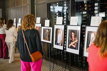 A woman in with blonde curly hair is admiring portraits framed in white mat and black frame placed on an easel displayed in the exhibit and reading the stories of the women in Journeys of women series in Minneapolis Minnesota by Portrait by Taru Studio, a headshot and personal branding photographer.
