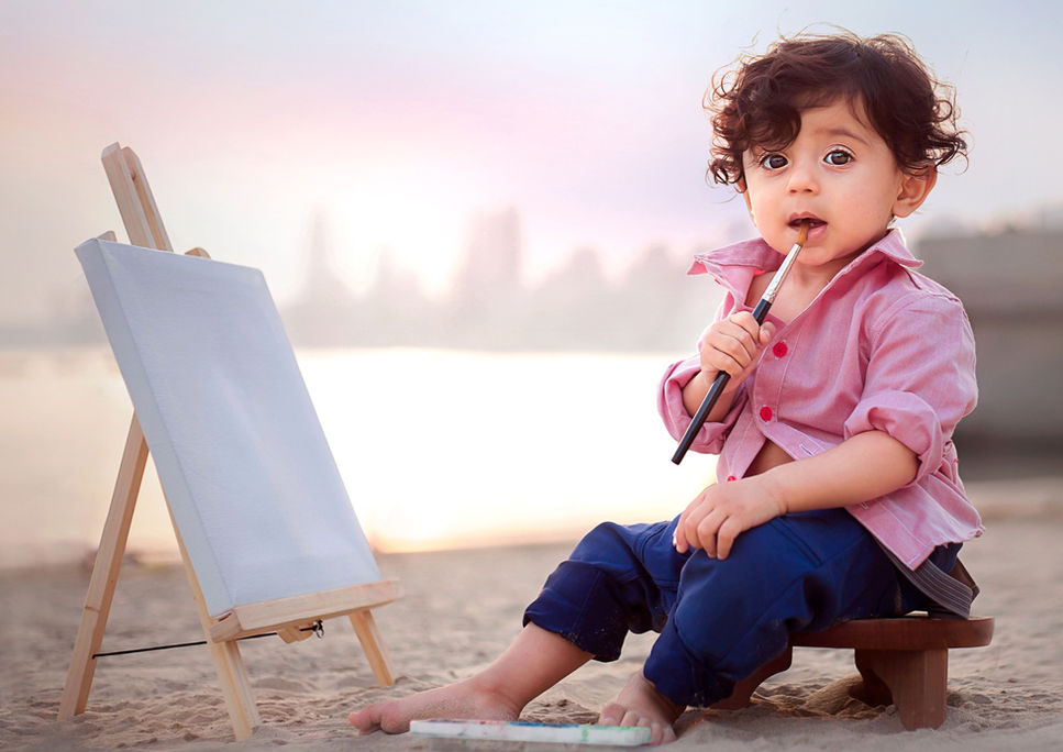 First birthday boy outdoor photoshoot by Taru Chhaya of portraitbytaru with a canvas on an easel and the baby holding paint brush on the beach with city skyline in the background in the photography shoot session