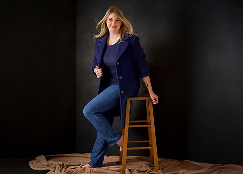 A woman wearing a blue woolen coat and blue denim jeans with blonde hair is looking at the camera with a smile at the photography studio for a Journeys of women portrait series by the headshot and personal branding photographer Taru Chhaya in Minneapolis Minnesota Midwest MN.