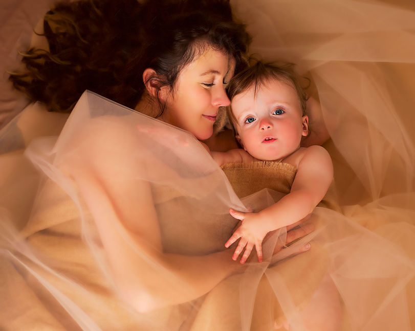 An indoor Studio motherhood session and first birthday photo shoot where the mother is looking and laying next to her one year old baby girl with blue eyes and golden blonde curly hair wrapped in a cashmere scarf and tulle for a Mom and me portrait by Taru, a Minneapolis based Midwest Minnesota photographer specializing in maternity, child, high school senior and family fine art photography