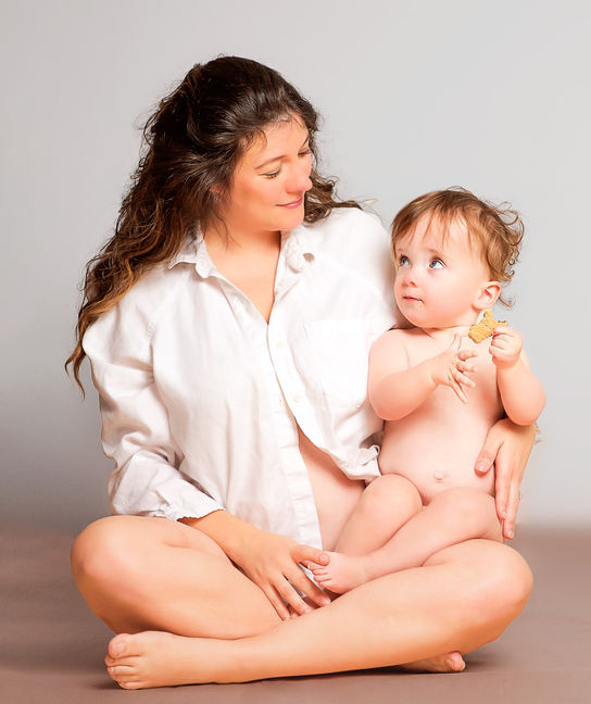 A one year old baby girl sitting on the lap of her mother is holding a cookie with her tiny fingers and the pregnant mom wearing a white shirt of her husband is looking at the daughter with golden curls and blue eyes for an indoor studio maternity mom and me first birthday photo shoot session by Portrait by Taru, a child, senior, family photographer in Midwest Twin Cities Minneapolis Minnesota MN