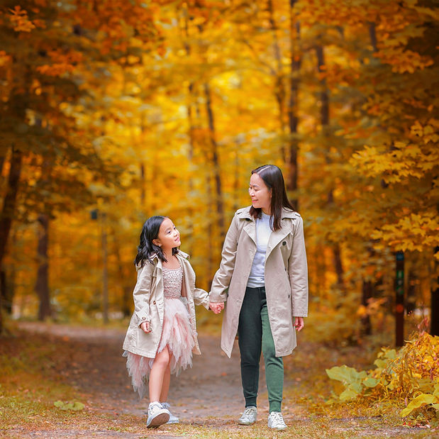A mother daughter picture where the tween girl wearing a trench coat on her favorite tulle dress is holding her mothers hand while the mom is looking at her child with all the love in her eyes among the best fall colors of beautiful yellow maple trees in Minnesota regional park for an outdoor birthday photo shoot by family photographer Portrait by Taru Studio in Minneapolis MN Midwest.