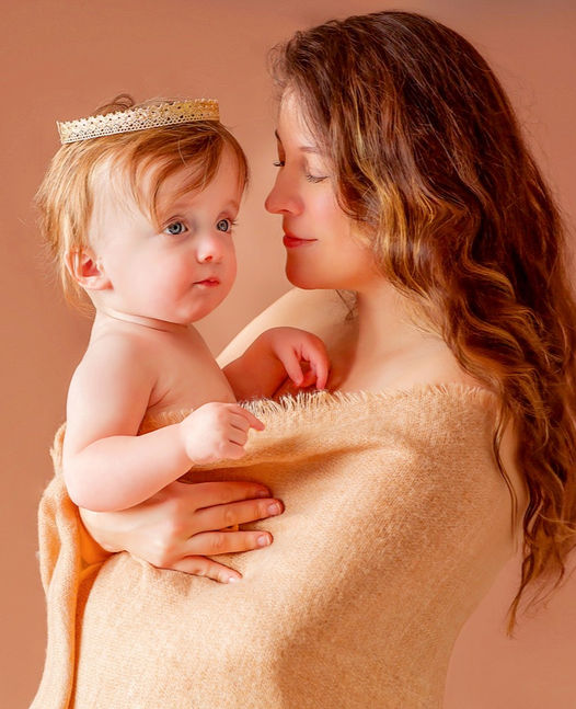 A one year old baby girl with blue eyes on her first birthday is wearing a lace gold crown on her red head while in the arms of her mother for a mom and me indoor motherhood studio photo shoot session by Portrait by Taru a Midwest Minneapolis based child family school senior and maternity photographer in Twin Cities MN