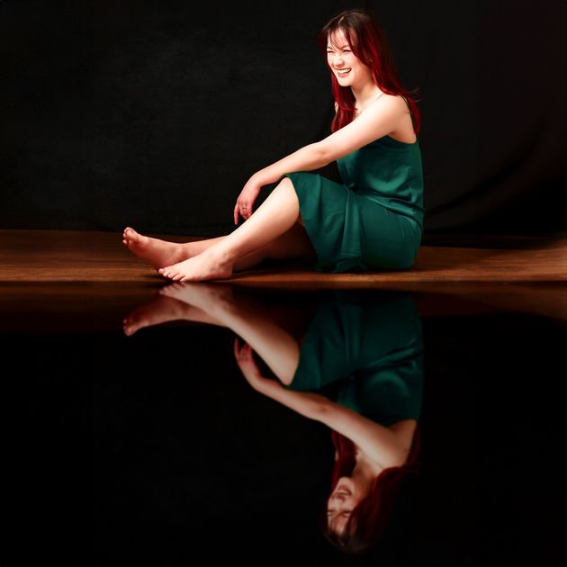 The empowering and transforming headshot branding photoshoot session of an asian American High School Senior girl sitting down on the floor in a green dress with her hand on her knee and laughing silly with a reflection of her laughter and herself nearby. The business entrepreneur women came in for her self-love and self-care photo shoot for her birthday gift at Portrait by Taru Studio, a maternity senior and family photographer in Plymouth Minnesota MN Midwest.