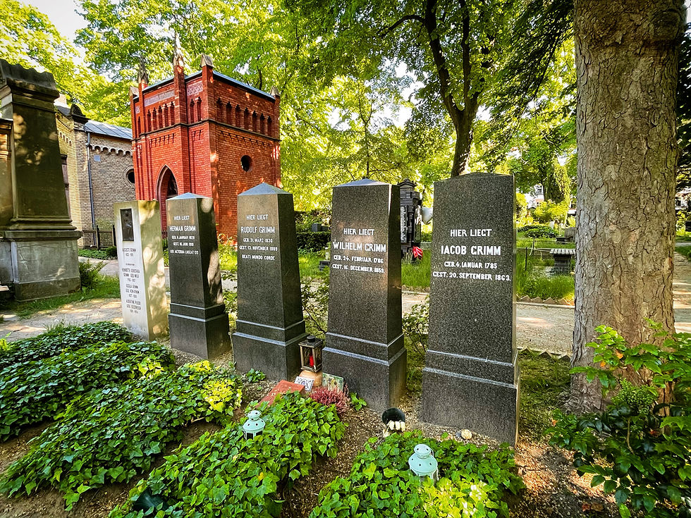 Grimm family graves in a leafy cemetery, with stone markers for Jacob, Wilhelm, Rudolf and Hermann Grimm and a red brick mausoleum.
