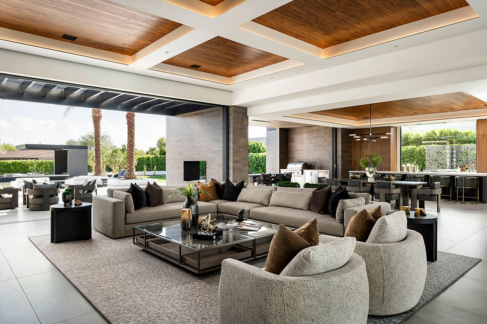 A bright, open living space with cozy neutral sofas and warm wood ceilings that flow seamlessly into an outdoor lounge and kitchen area surrounded by palm trees and greenery.