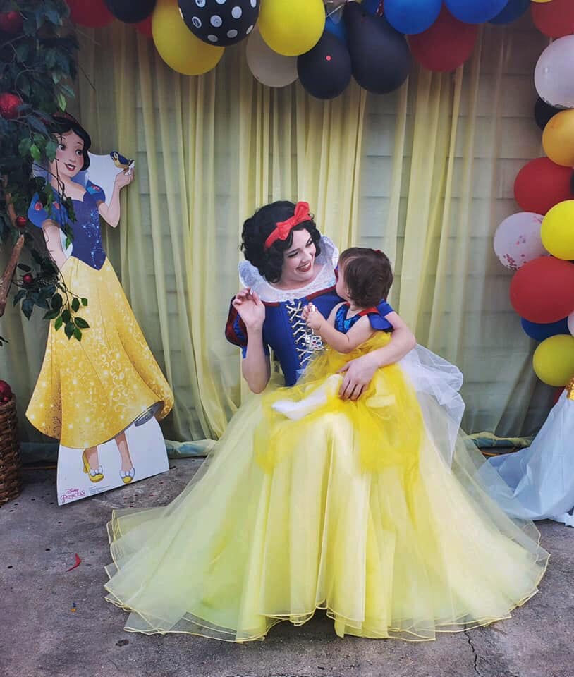A person dressed as Snow White holds a child in a matching outfit, smiling. They're surrounded by colorful balloons and a Snow White cutout.