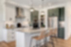 Renova's showhome features a modern kitchen with white and sage green cabinets, stainless steel appliances, marble backsplash, island with sink, pendant lights, and wicker stools.