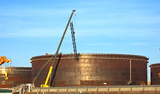 Aerial view of a large oil storage tank under construction, with a spherical stairway spiraling upwards around its exterior. A construction crane stands beside the tank, indicating on going development