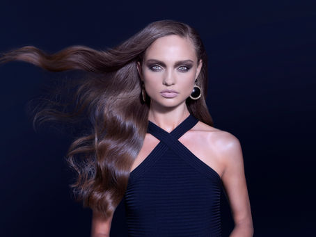 Woman with flowing brown hair, smokey eye makeup, and hoop earrings in a dark setting. Wearing a criss-cross black top, serious expression.