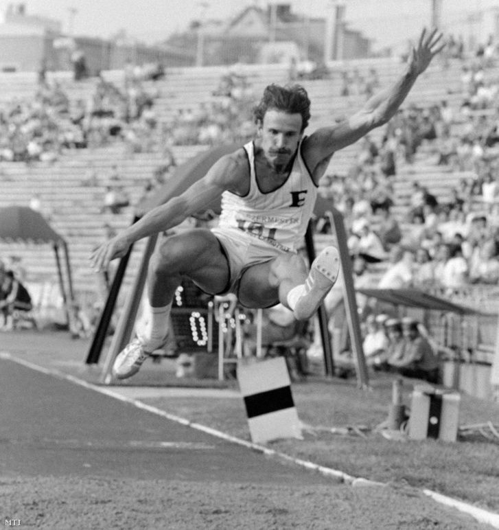 fekete-fehér fotó az 1980-as évekből, egy bajszos férfi távolugró atlétaként, fehér mezben épp ugrás közben repül a homokos pálya felett egy versenyen, a háttérben lelátó a közönséggel