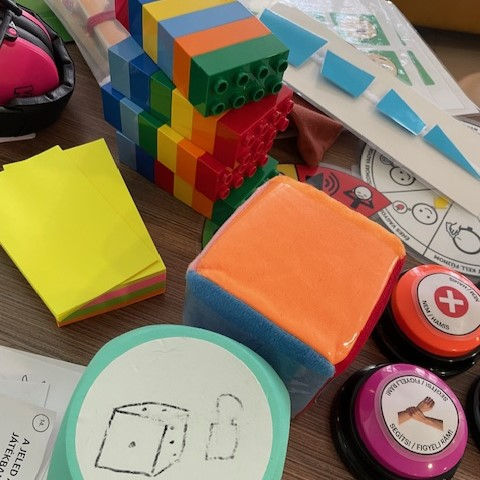 Színes eszközök egy asztalon: Duplo kockák, puha dobókocka, post-it papírok, nyomtatott és papírból kivágott minták