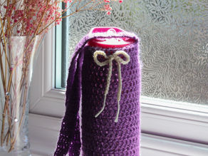 Purple crochet water bottle holder with a white bow, on windowsill. Floral frost on glass, pink flowers in vase, sunlight streaming in.
