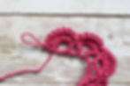 Pink crochet pattern in progress on a wooden background, with loops and stitches visible. The yarn contrasts with the white wood texture.