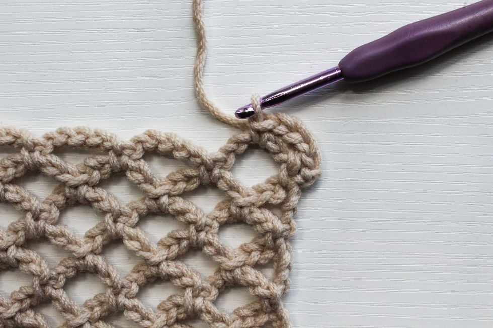 Crochet diamond mesh in beige yarn in progress. A purple crochet hook is looped through yarn on a white textured surface.