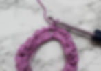 Close-up of a purple crochet scrunchie in progress with a silver hook on a marble-patterned background. Yarn looped through hook.