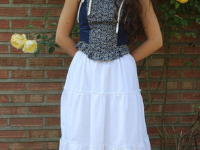 Woman in a blue floral top and white skirt stands by a brick wall, surrounded by yellow roses, looking contemplative.