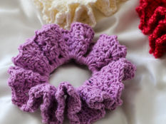 Crocheted scrunchies in purple, white, and red sit on a silky, shiny white fabric background.