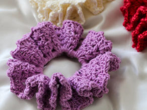 Crocheted scrunchies in purple, white, and red sit on a silky, shiny white fabric background.