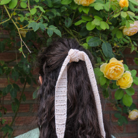 Back of a woman with long dark hair tied by a cream crocheted ribbon, standing before yellow roses and green foliage against a brick wall.