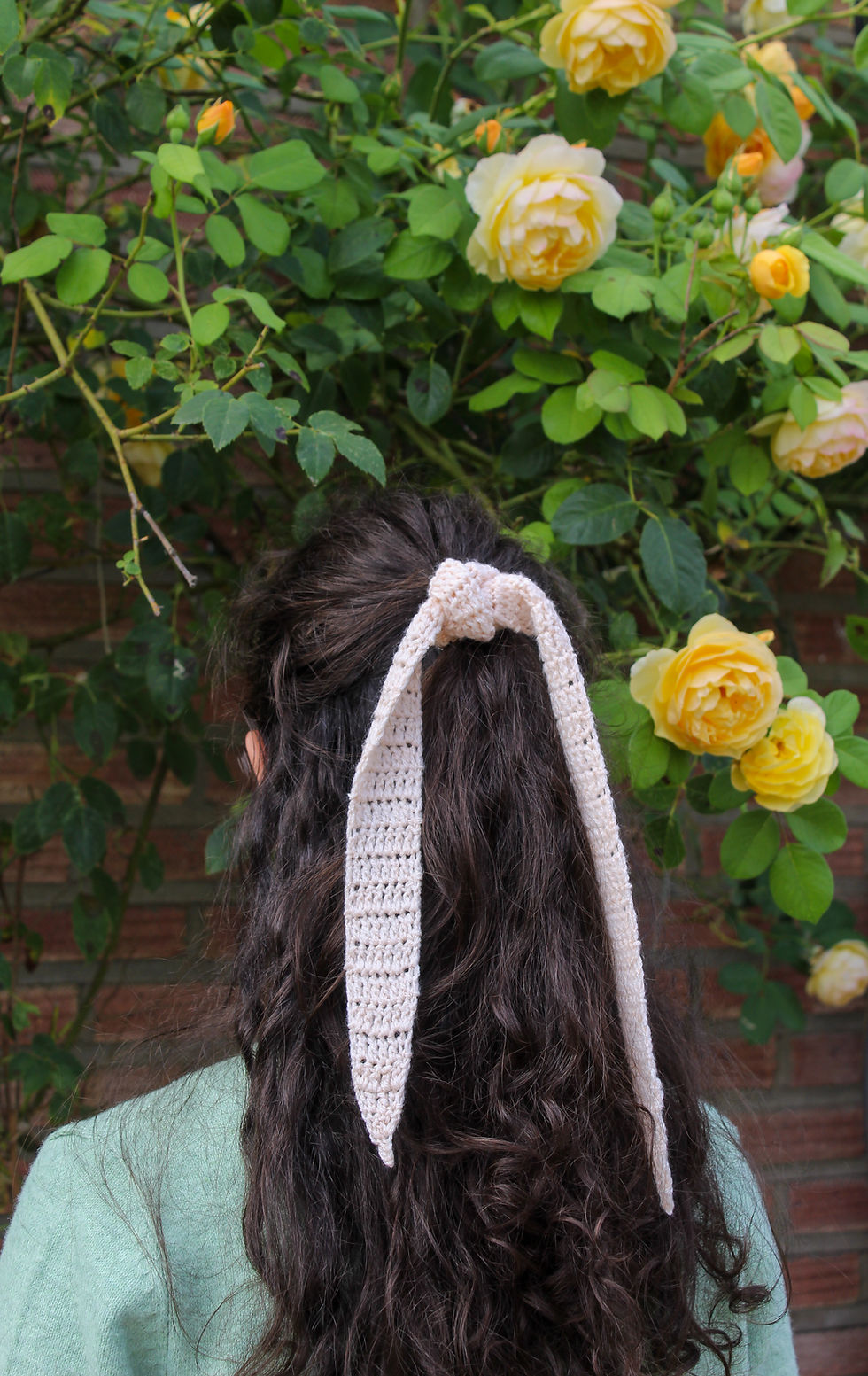 Back of a woman with long dark hair tied by a cream crocheted ribbon, standing before yellow roses and green foliage against a brick wall.