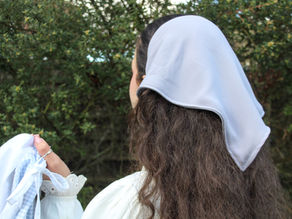 Woman with long hair wearing a gray headscarf, holding a light blue and white cloth. Lush green foliage in the blurred background.